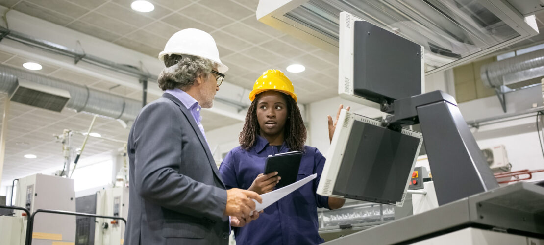 Black female factory worker and her male boss standing at industrial machine and talking. Low angle. Production process or machinery concept